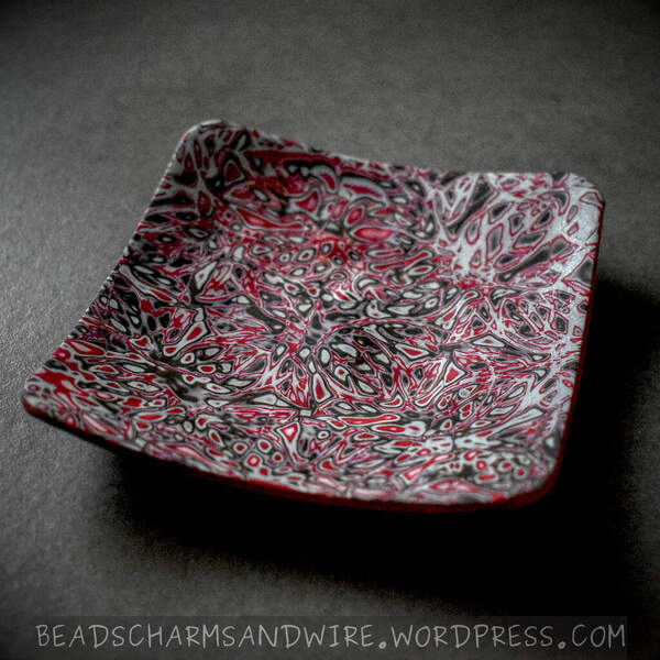 Polymer clay ring dish with a rounded square shape and a curved surface raised higher at its corners. The ring dish is completely covered with a random abstract pattern using the mokume-gane technique in red, gray, white, and black.