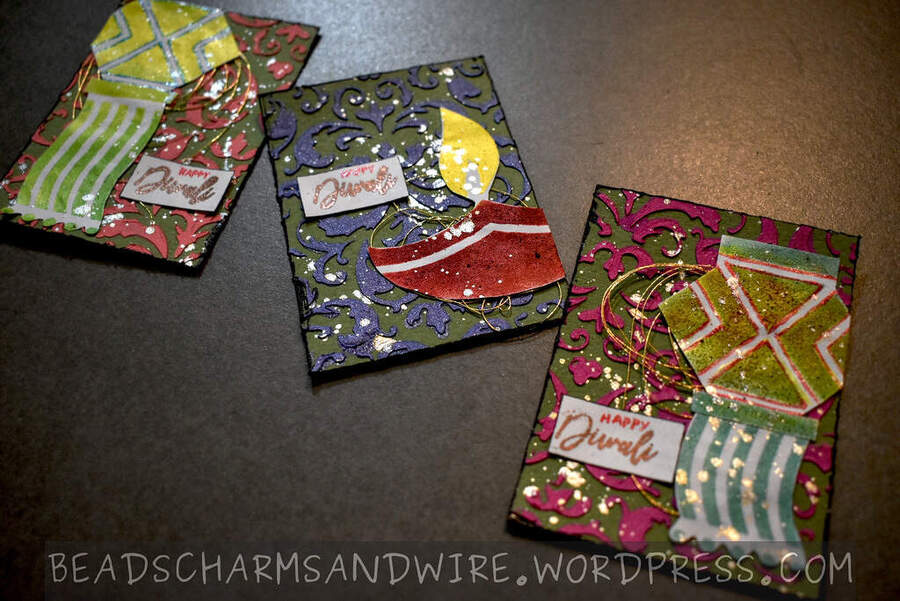 A trio of mixed media ATCs with colored, textured backgrounds, and Diwali motifs like diyas and hanging lamps in the foreground. The text 'Happy Diwali' is attached alongside the motifs.