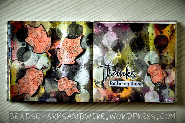 An art journal spread across two square pages, with a bokeh-like background of black, white, and colorful circles, and splatters. The left page shows four orange leaves, large and small; the right page includes the words 'Thanks for being there' and a small orange leaf to the side.