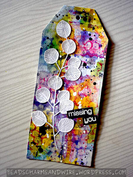 A mixed media tag with a colorful bubbly background, and a focal white die-cut of eucalyptus leaves. The words 'missing you' are embossed at an angle in the lower half of the tag.