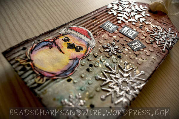 A Christmas-themed mixed media tag made of corrugated cardboard, with cardboard ridges. stenciled texture. and sparkly snowflakes in the background. The focal element is a little bird wearing a Santa hat. The words 'Love', 'Peace', and 'Joy' are stamped in glitter on black background above the bird.