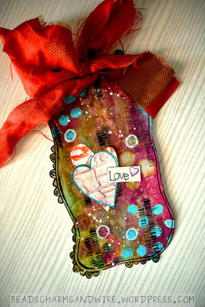 A wavy mixed media tag with protruding doodles on some parts of its edges. The background is pink, brick, and green; with teal, black, and white marks and white splatters. Two textured, overlaid, asymmetrical hearts form the focus, and over them is a sentiment 'Love'. There is a red fabric bow at the top of the tag.