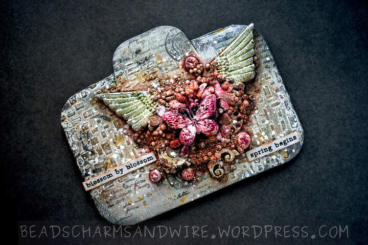 A mixed media assemblage project on a landscape-oriented tag base. The background is filled with texture from embossed and stamped areas. The focal objects are heaped one over another, with wings at the bottom, followed by flowers, steampunk gears and screws, and a topmost butterfly visible amongst a lot of small round pebbles.