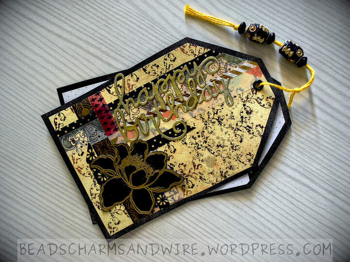 A mixed media tag duo with a washi-tape-covered, primarily yellow background. In the bottom-right corner is a black flower embossed in gold, and along the upper left edge is a large gold 'happy birthday' sentiment. The tags are joined with a cotton cord that has gaudy beads strung on it.
