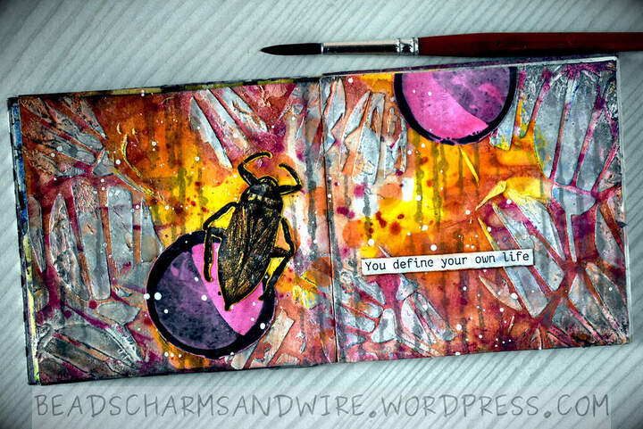 A colorful yellow-orange double-page spread on a 10x10 art journal page, with whitish textured flowers and vertical squiggles in the background. A couple of black-outlined circles, colored in pink and a lighter black are spread out over the pages. Over the bottom circle on the left page is a black beetle with a yellow sheen. A strip of paper that reads "You define your own life" is along the lower right page.